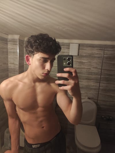 A shirtless young man with curly hair takes a selfie in a bathroom. He holds a dark-colored smartphone to capture the image, tilting his head slightly downward. The bathroom features gray-toned tiled walls, a toilet, and visible overhead lighting casting shadows. His physique reveals defined pectoral and bicep muscles. He wears dark-colored bottoms.