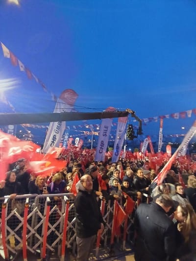 The image shows a large crowd of people gathered in an outdoor space in Istanbul, Türkiye. The location of the crowd is near the coordinates 41.084, 28.678, which corresponds to a street near Taksim Square. The crowd is standing behind a metal barrier and is holding red flags with white text that says "CHP" and "CHP SILE". The crowd is facing a stage that is not visible in the image. In the foreground of the image are a large number of people wearing jackets, coats, and hats. The crowd is diverse in age and gender, with many families present. The crowd appears to be waiting for an event to begin. The sky in the image is blue with some clouds. The event is likely a political rally or a campaign stop for the CHP (Republican People's Party) political party.