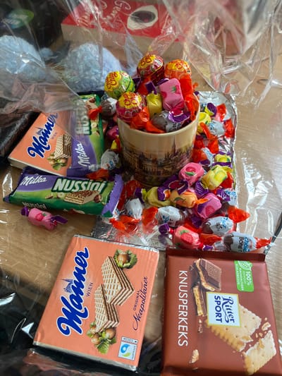 A mug filled with various wrapped candies and lollipops sits on a silver tray, surrounded by additional loose candies. The tray is partially covered by plastic wrap.  The mug is printed with an image of the city of Wien, Austria, with the visible details of buildings, a river, and bridges. The tray contains several boxes and wrappers of various Austrian chocolate brands, including Manner, Nussini, Ritter Sport, and Knusperkeks. The image was likely taken in a residential setting, in the city of Wien, Austria, as per the coordinates provided (48.201, 16.339).  The composition suggests a celebratory occasion, possibly a birthday or a gift exchange. The overall scene is colorful and suggests a joyous atmosphere.