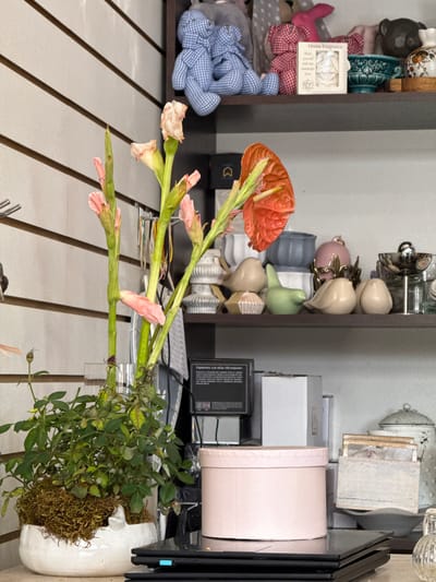 The image depicts a still life scene indoors, likely in a home or shop setting in Sunzha, Russia. The primary subjects are decorative objects and plants. In the foreground, an orange anthurium flower with green stems and buds is prominent. To the right, a potted plant with green leaves and brown moss surrounding its base is also visible. Behind these floral elements, a wooden surface with light-colored slats and dark lines runs horizontally.

Further back, shelves are filled with various decorative items. These include ceramic figurines of birds and tulips, a pink ceramic pot, and what appear to be decorative vases or pitchers. There are also several boxes, some white and some light pink, stacked on a shelf. A black electronic device with some visible text is also present. In the upper left portion of the image, more decorative items are visible, including what looks like ceramic dishes and a small white box with text on it, possibly a product label for "Home Fragrance."

The scene is brightly lit, suggesting it might be daytime, and there are no immediate indications of weather conditions. The overall impression is one of a curated display of decorative items, possibly for sale or as personal ornamentation. No people are visible in the image. The text on the black device is partially visible and appears to be in Cyrillic script, consistent with the provided location context.