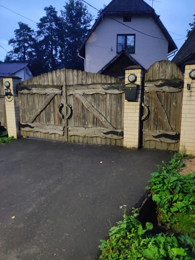 This image shows a set of double wooden gates in front of a house in the city of Alabino, Russia. The gates are made of vertical wooden planks with decorative diagonal braces and arched metal accents. The gates are flanked by brick pillars, one of which has a black mailbox attached. A round light fixture is mounted on top of each pillar. A house with a dark roof and light-colored siding is visible behind the gates. Trees are silhouetted against the twilight sky. The ground in front of the gates is paved with asphalt, and there is lush green vegetation growing along the edge of the asphalt. The overall impression is one of a private residence with a rustic and somewhat imposing entrance.