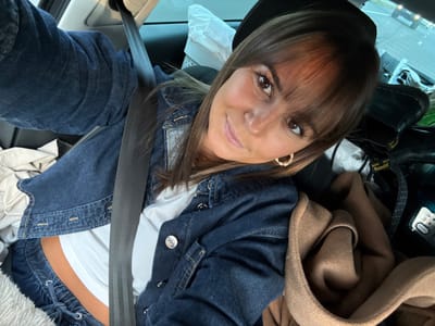 The scene depicts a female individual inside a vehicle, reportedly situated in Bourg-lès-Valence, France. The subject, possessing brown hair with bangs, light brown eyes, and a tanned skin tone, is observed wearing a dark blue denim jacket, a white cropped undershirt exposing the midriff, and dark blue drawstring pants. Gold-colored, circular earrings adorn her earlobes. A black vehicular seatbelt extends diagonally from the upper left across her torso to the lower right. A dark-colored cap or hat is partially visible on the subject's head. The vehicle's interior background includes white fabric on the left side, a folded brown textile object (possibly a coat or blanket) on the right, and a pair of black boots with distinct yellow stitching, indicative of a brand such as 'Doc Martens,' positioned near the rear right window. A blurred exterior environment, suggestive of a road or highway, is visible through the vehicle's windows.