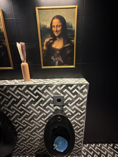 The image shows a bathroom scene with a black and white patterned tiled wall and floor. A black toilet bowl with a blue cleaning disc is visible. On the black tiled wall to the left, there is a framed picture of the Mona Lisa. A decorative vase with white and brown plumes is positioned between the Mona Lisa picture and another framed picture below. The location is Ulm, Germany.