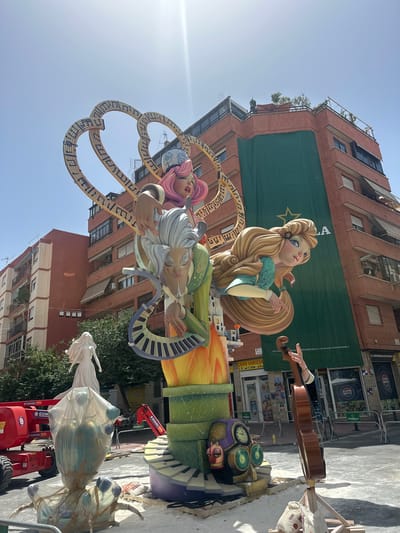 This image captures a vibrant street scene in Alicante, Spain, likely during the construction or setup phase of a festival or art installation. The primary subjects are elaborate, large-scale figures that appear to be part of a "falla," a traditional Valencian monument.

In the foreground, a woman's hand is seen playing a cello, suggesting a musical theme. Behind this, a stylized sculpture depicts several figures, including a blonde woman with flowing hair and two older figures with musical instruments integrated into their design, such as a keyboard. These figures are rendered in bright colors and dynamic poses, hinting at a festive and celebratory atmosphere. A large heart-shaped structure adorned with musical notes further emphasizes the musical motif.

The background features a multi-story brick building, partially covered by a large green banner with a star emblem. Various shops and storefronts are visible at street level, with signs in Spanish, including "SUPERMERCADO ESPAÑA," "ESTRELLA," and "PEPSI," providing clear location cues. A red cherry picker, labeled "GOMARIZ," is present, indicating ongoing work or assembly.

The lighting suggests it is daytime, with a clear sky. The overall scene is one of anticipation and artistic creation, with the detailed sculptures being the focal point against the urban backdrop. The presence of construction equipment and the partially completed nature of some elements point to an event that is either in preparation or underway.