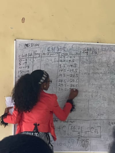 The image captures an indoor scene in Uturu, Nigeria, specifically a classroom. A female individual, positioned with her back partially turned toward the viewer, stands facing a large white dry-erase board. She has long, dark, wavy hair pulled back, secured with a black and white patterned headband, and wears glasses. Her attire includes a coral-colored blazer with black ruffled details on the cuffs, and a black and white striped skirt. She holds a dark marker in her right hand, actively writing numerical values and text on the whiteboard, while her left hand holds several white sheets of paper.

The whiteboard displays statistical calculations related to finding the median. Visible headers include "MEDIAN," "Class Int.," "freq," "Mid. point," "class boundary," "freq," and "FX." Specific class intervals like "0-4," "5-9," "10-14," and corresponding class boundaries such as "0.5-4.5," "4.5-9.5" are written. A formula for the median, "L + C [(Ef + 1)/2] - Cfb," is partially visible at the bottom. The board exhibits signs of previous use, with multiple layers of erased and smudged writing. The wall behind the whiteboard is a pale yellow color. A small, reddish, clustered object is affixed to the upper left portion of the wall. In the lower foreground, a dark, out-of-focus shape indicates the presence of another individual, likely a student.