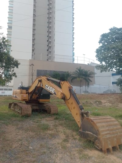 An outdoor, medium-angle, eye-level shot captures a yellow CASE excavator parked on a patch of dirt and grass in front of a tall, modern, white building in Cuiabá, Brazil. The excavator is positioned in the foreground, with its tracks resting on the ground and its bucket at an angle, partially lowered. The building, which appears to be an apartment or office complex, dominates the background with its numerous balconies and windows. Trees frame the scene on the left and right, and a few palm trees are visible near the building's base. Power lines are strung between poles in the middle ground. The sky is overcast and bright, suggesting a daytime scene, possibly with humid weather typical of Cuiabá. There are no people visible in the image.