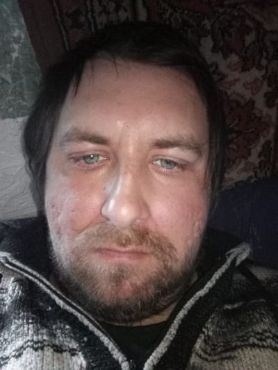 The image shows a close-up of a middle-aged man's face. He has light blue eyes, dark hair, and a beard with stubble. There are visible skin imperfections on his cheeks. He is wearing a black and white patterned sweater or jacket. The background behind him is a blurry mix of what appears to be patterned wall coverings. There is no evidence that can prove that the image was taken in Parabel, Russia.
