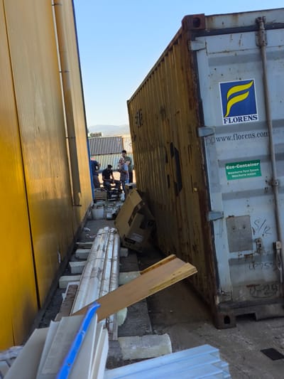 The image depicts an outdoor scene in the Municipal Unit of Inofyta, Greece, likely a port or industrial area, with a bright blue sky overhead. In the foreground, a tall, ribbed yellow wall occupies the left side of the frame, casting a shadow. To its right, a large, weathered shipping container is positioned, its side showing rust and graffiti, including the "FLORENS" logo and "www.florens.com". A smaller green sticker reads "Eco-Container Waterborne Paint System Waterborne sealant". Several people are gathered near the shipping container, some appearing to be working or observing. Among the objects present are wooden planks, bundles of materials wrapped in plastic, and what appears to be a blue tool or pipe. In the background, more shipping containers and industrial structures are visible, along with distant hills.