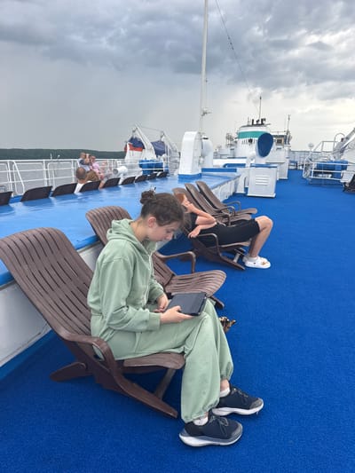 On the deck of a ferry with a blue carpet, a young woman in a light green tracksuit sits in a brown lounge chair, looking at her tablet. She is wearing dark sneakers and white socks. Behind her, another person, possibly a teenager, in a black outfit is also sitting in a lounge chair. In the background, other passengers are visible sitting and standing, and a part of the ferry's structure with lifeboats and other equipment is seen. The sky is cloudy. The location is Ikskoe Ust'e, Russia.
