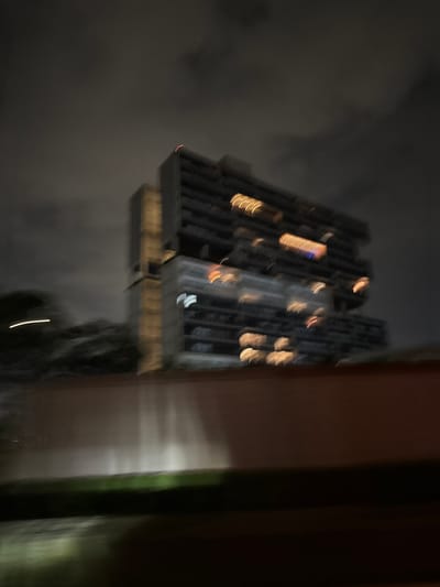 The image shows a tall building with numerous windows lit up at night. The building is dark gray with several illuminated sections, suggesting residential or office spaces. The surrounding environment appears cloudy and dark. There are elements of a structure or wall on the right side, which suggests the photo was taken from inside a building. There is no indication of the location being Santa Clara Ocoyucan, Mexico, or any specific identifiable landmarks or signs.