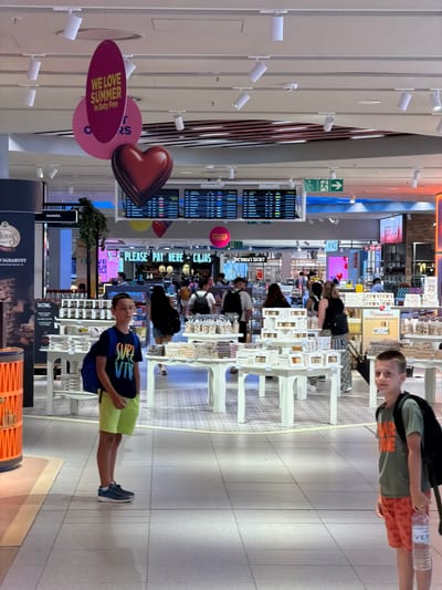 In a well-lit, indoor setting resembling a duty-free shopping area, likely an airport, two young boys are the primary subjects. One boy, in the foreground, wears a blue and green t-shirt and light green shorts, holding a bottle of water. He stands on a tiled floor, looking towards the camera with a neutral expression. Behind him, shelves stocked with various products, including what appears to be food items and cosmetics, are visible. Signs in English and Spanish are present, such as "WE LOVE SUMMER in Duty Free," "VICTORIA'S SECRET," "SANGRIA," and "TASTING."
In the background, another boy, wearing a green t-shirt and orange shorts, is also seen holding a bottle of water. He is looking away from the camera. Other shoppers and staff are present, browsing or attending to displays. The overall atmosphere suggests a busy commercial environment. The lighting is bright and artificial, indicating it is daytime. The presence of duty-free advertising suggests this is likely a travel hub.