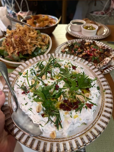 A close-up, overhead shot shows a rustic, creamy dip in a decorative ceramic bowl, garnished with herbs, nuts, and red flecks. A spoon rests in the dip, and a woman's hand is visible on the left side of the frame. In the background, other dishes of food are partially visible, along with a blurred restaurant setting. The scene is likely in Abu Dhabi, United Arab Emirates.