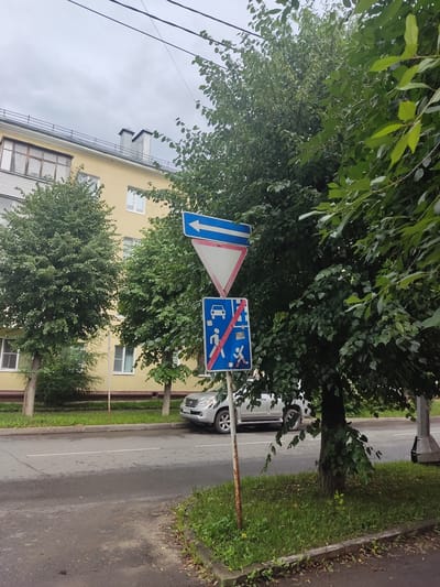 This image depicts a street scene in Serpukhov, Russia, likely during the daytime under overcast skies. The foreground is dominated by lush green trees, their leaves forming a dense canopy that partially obscures the view of the background. A yellow building with several windows is visible behind the trees, suggesting a residential or commercial area.
Several traffic signs are prominently displayed. One sign is a blue rectangle with a red diagonal line across a depiction of a car and a person walking, indicating a prohibition or restriction. Another sign, a white triangle with a red border and a blue arrow pointing downwards, likely signifies a directional instruction or warning. A metal pole, showing signs of rust, supports these signs.
The street itself is paved and appears wet, suggesting recent rain. A silver car is parked on the side of the road, and a grassy verge runs alongside the street, lined with small plants and fallen leaves. Power lines are also visible stretching across the sky, indicating an urban or semi-urban environment. The overall impression is that of a quiet, ordinary day in a tree-lined street, with the traffic signs offering context to the specific regulations of the area.