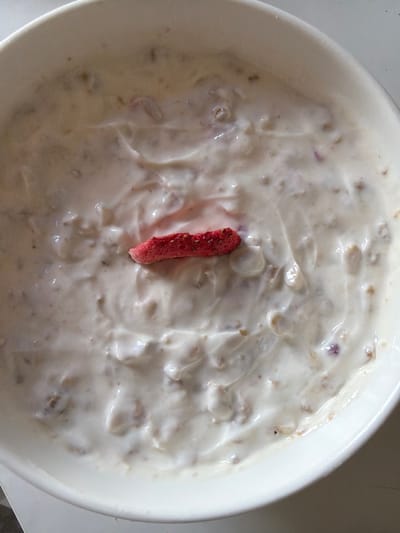 The image is an overhead shot of a white bowl containing a creamy, off-white substance, likely yogurt or a similar dairy product, mixed with visible pieces of what appear to be oats or granola. A single, small, bright red, freeze-dried strawberry slice is positioned centrally on top of the mixture. The background is out of focus, with only hints of a light-colored surface, possibly a table. There are no discernible landmarks or features that would allow for location identification within Boxtel, Netherlands. The scene is static and appears to be a close-up of food.