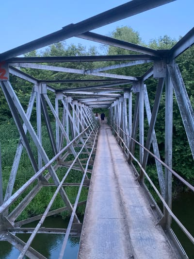 This is a photograph of a pedestrian bridge in the city of Yuryatino, Russia. The bridge is constructed of grey metal beams and girders, forming a lattice-like structure. The walkway is made of metal plates. Two people are visible walking away from the camera on the bridge, one with dark clothing and the other with lighter clothing. The bridge spans a body of water, which appears to be a river or canal, and is surrounded by lush green trees and foliage. The sky above is a clear, pale blue, suggesting it is either early morning or late evening. There is some graffiti on the metal structure.