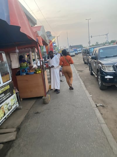 A street scene shows a row of cars parked along the roadside. A fruit stand with an awning displays oranges and other produce. Two individuals walk along the sidewalk, one in a white robe and the other in an orange shirt and beige pants. A sign with food is visible. The sky appears overcast, with streetlights and overhead wires lining the street.