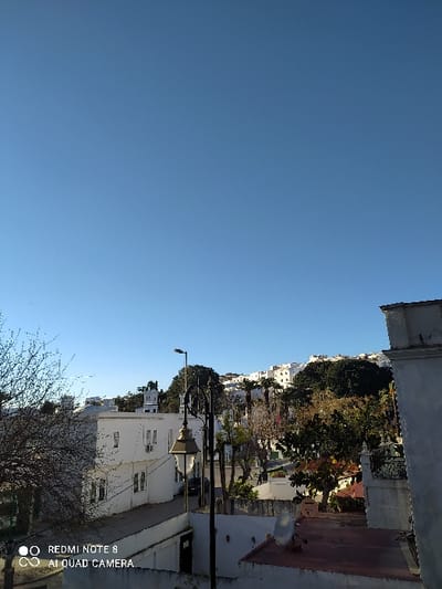 A view captures a city street nestled between white buildings. Sunlight dapples the trees lining the road. The sky is clear and blue. An antenna sits on a roof of one of the buildings. The camera's watermark is visible in the corner.