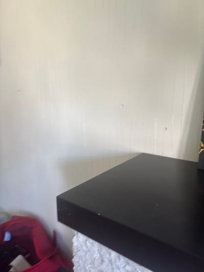 The image shows a corner of a room with a black shelf or table in the foreground. The wall behind is light-colored and appears to have some vertical texture. A red bag is visible to the left of the shelf. There are no people or any other discernible objects in the scene. The location is not determinable from the image content.