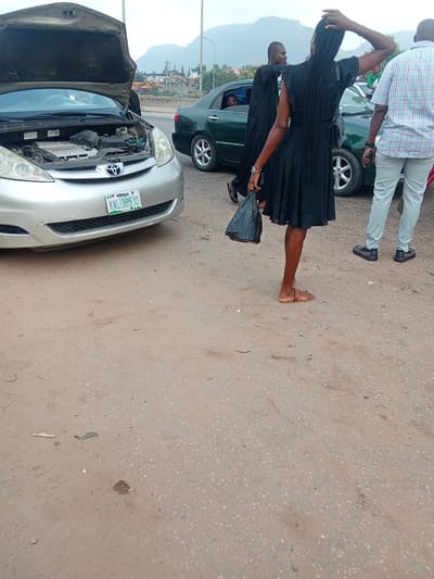 In Kagini, Nigeria, on an unpaved road, a silver Toyota Sienna minivan with its hood open is parked. The minivan's engine is visible. A dark-colored sedan is parked behind the minivan, and a green sedan is to the right of the minivan. A man in a checkered shirt and grey pants stands to the right of the scene. A woman in a black dress with braided hair is standing between the two sedans, holding a black plastic bag. Her left hand is on her head, and her right hand is by her side. Another man in a black robe is partially visible in the background, looking towards the woman. The environment is dusty with sparse vegetation and a mountainous backdrop.