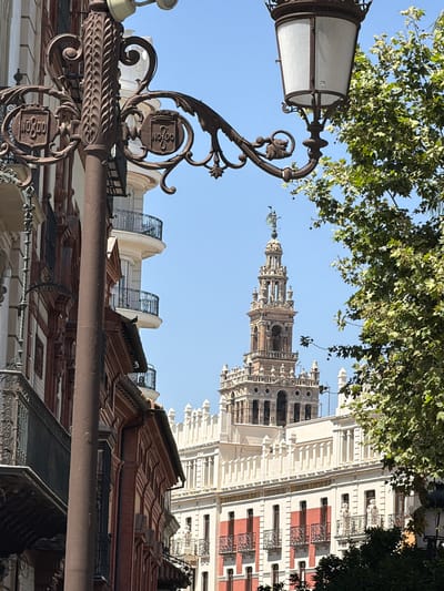 The image captures a street scene in Seville, Spain, with a prominent ornate lamppost in the foreground. Behind the lamppost, a tall, intricately designed tower, likely the Giralda tower, rises against a clear blue sky. The tower is flanked by buildings with traditional Spanish architecture, featuring balconies with wrought-iron railings and decorative facades. Green foliage from trees partially obscures the view of the buildings and the tower. The scene conveys a sense of historic European urban landscape on a sunny day.