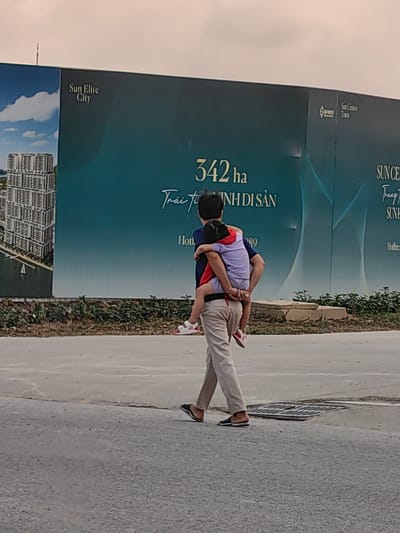 An adult, likely male, is depicted walking away from the camera, carrying a child piggyback on a paved road. The adult wears a dark blue shirt, light beige trousers, and dark slippers. The child, dressed in a light purple top and red shorts with white sandals, clings to the adult's back.
The background features a large, dark blue-green billboard advertising a real estate development. On the left panel, "Sun Elite City" is visible above an illustration of high-rise buildings. The central section displays large white text: "342 ha" (hectares) and "Trái tim DI SẢN," meaning "Heart of Heritage." A partial "Hotline" number, ending in "89," is also visible. Further right, the billboard shows "Sun Centro Town," with additional fragmented text "SUN C" and "SUN E."
The scene is set outdoors during the daytime, under an overcast sky. Sparse greenery is present at the base of the billboard, and a manhole cover is on the right side of the road. The visible Vietnamese text and real estate advertisements suggest the location is a developing area in Bai Chay Ward, Vietnam. The image captures a quiet moment of familial care as the adult carries the child.