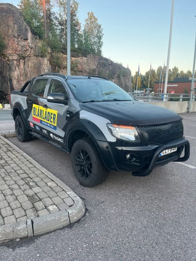 A gray Ford Ranger pickup truck with "BLAKLADER WORKWEAR" branding is parked on asphalt next to a cobblestone sidewalk in Vantaa, Finland. The truck has a black bull bar on the front and black fender flares. In the background, a rocky cliff face with trees is visible, along with a McDonald's restaurant and several flags. The sky is clear and blue. The license plate on the truck reads "CLT 846".