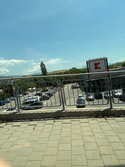 The image captures a view from an elevated position, likely a balcony or rooftop, overlooking a parking lot with the Kaufland store sign visible in the background. Cars are parked in rows within the lot, and a metal railing separates the vantage point from the parking area. Beyond the parking lot, there is a landscape with trees and vegetation, partially obscured by the railing. A clear blue sky with a few clouds is visible. The paved surface of the balcony or rooftop is in the foreground. The location is identified as Sandanski, Bulgaria, based on the Kaufland sign and general context.