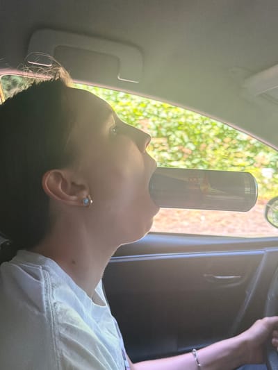 A medium shot captures a person's profile as they drink from a Red Bull can in a car. The person is wearing a white t-shirt and a small earring in their left ear. The car's interior is visible, with a sun visor overhead and the dashboard and door panel on the right. Green foliage and trees are seen through the car window. The image was taken in Seaside, United States.