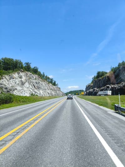 The image shows a highway in Papineauville, Canada, with a bright blue sky and scattered clouds. On the left side of the road, a rocky cliff face is covered with lush green trees. On the right side, another rocky cliff face is visible, with a white truck parked alongside it. The truck has "Nadeau" written on its side. A silver SUV is driving on the highway ahead, and yellow road signs are visible in the distance. The road is paved with asphalt and has double yellow lines dividing the lanes. The overall scene depicts a scenic drive through a natural landscape on a clear day.