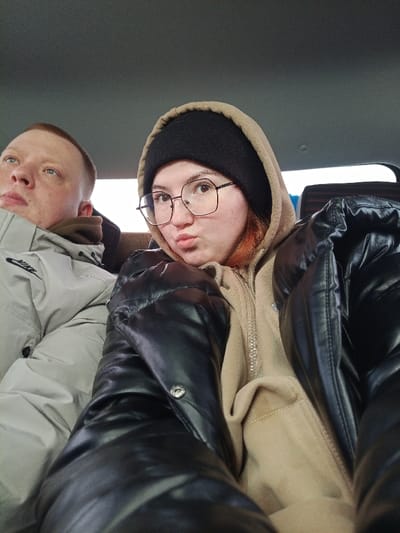 The image depicts two individuals seated within the interior of a vehicle. The specific location in Kueda, Russia, cannot be determined from the visual information provided.
In the foreground, positioned to the right, is a female subject with light skin, reddish-brown hair visible under a black knit beanie, and dark hexagonal-framed eyeglasses. She wears a tan hooded sweatshirt, zipped, underneath a shiny black puffer jacket. Her lips are pursed, and she looks directly at the camera.
To her left and slightly behind her, a male subject with light skin and short light-colored hair is visible. He wears a light grey puffer jacket, with a partial "NIKE" logo visible on the left sleeve, and a brown collar or garment under his jacket. He is looking upwards and to his left, away from the camera.
The vehicle's interior features a grey fabric headliner. A dark, possibly striped, pattern on a seat or wall is faintly visible behind the female's right shoulder. Both individuals appear to be passengers. The scene is illuminated by ambient light, suggesting daytime.