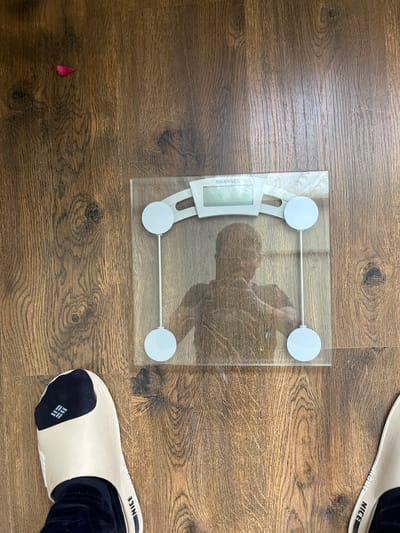 A clear glass digital scale with a white display screen showing the brand "MAXWELL" is placed on a wooden floor. A person's reflection is visible on the scale's surface, showing their upper body and head. A single pink flower petal lies on the floor to the left of the scale. The bottom of the frame shows a person's feet wearing beige slides over dark socks. The floor appears to be laminate or hardwood, with visible wood grain. The scene is indoors, likely a home, given the flooring and personal items. There is no information or context to suggest this scene is located in Berd, Armenia; it is an indoor domestic setting.