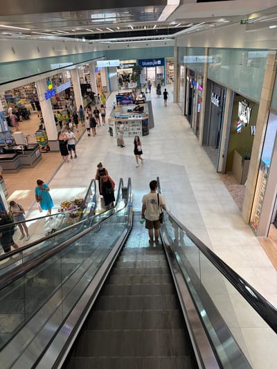 The image depicts an interior view of a modern shopping mall, likely in Olomouc, Czechia, characterized by its bright lighting, tiled floors, and multiple retail storefronts. In the foreground, two escalators descend, with people using them. On the left escalator, a person in a blue dress and a woman in a black dress are descending, while on the right escalator, a man wearing a white t-shirt and khaki shorts is going down.

Further into the mall, a variety of shops are visible. On the left side, an "Albert Hypermarket" is prominent with many shoppers. To the right, there are storefronts labeled "Bluestyle" and "Cistiarna." In the mid-ground, several people are walking and interacting. Towards the right side of the mall, a barber shop with a striped pole logo and the name "THE ORIGINAL WALK-IN BARBER" is visible. The overall environment is clean, spacious, and well-lit, suggesting a typical commercial setting.