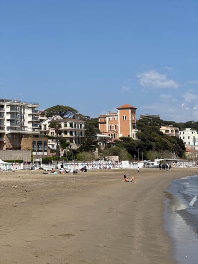 The image depicts a sunny beach scene in Anzio, Italy. In the foreground, a sandy beach stretches towards the ocean's edge, where gentle waves are visible. A person is lying on the sand, seemingly sunbathing. Numerous beach umbrellas and chairs line the edge of the beach near the buildings. Behind the beach, buildings rise up the hillside. The architecture varies, with a mix of older, brick structures and more modern white buildings. The sky above is a clear blue with a few scattered clouds.