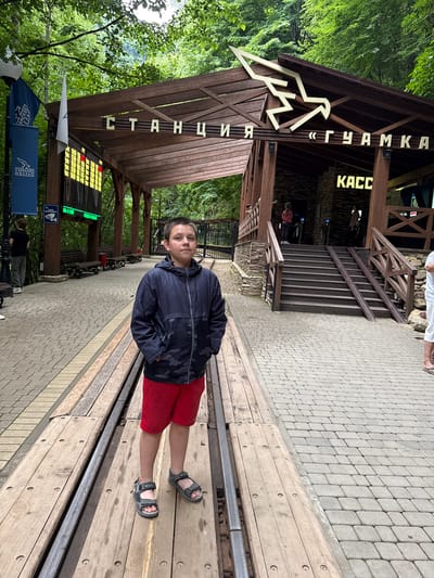 A young boy stands on a wooden platform next to train tracks at the Guamka station in Guamka, Russia. The boy is wearing a dark blue camouflage-patterned jacket, red shorts, and grey sandals. Behind him is a wooden structure with signs in Cyrillic script. One sign reads "СТАНЦИЯ «ГУАМКА»" (Station "Guamka"), and another smaller sign reads "КАСС" (Cashier/Box Office). A large electronic display board showing numbers is visible to the left. The station is surrounded by lush green trees, indicating it is an outdoor, natural setting. Several benches are present on the platform, and a paved walkway with cobblestones runs alongside the tracks. Other people are partially visible in the background near the station building and on the stairs leading up to it. The weather appears to be overcast.
