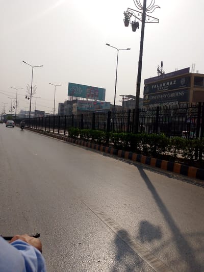 The image captures a street scene in Peshawar City Tehsil, Pakistan, under a bright, overcast sky. In the foreground, a person's hand and arm, wearing a blue shirt, are visible, suggesting the photo is taken from the perspective of someone on a motorbike or bicycle. The road surface is asphalt, with a distinct painted lane marker.
The middle ground shows a multi-lane road with light traffic, including a white car and a motorcycle. A black metal fence lines the right side of the road, behind which are manicured green bushes. Beyond the fence, several buildings are visible. One prominent building displays signage for "Falaknaz Group" and "Discovery Gardens," along with what appears to be a "Marks & Spencer" advertisement. Another billboard advertises "Cold Coffee Kick" with images of Nescafe products. Streetlights, some with decorative elements and hanging flower pots, are positioned along the roadside.
The background is dominated by the hazy sky and the outlines of further buildings and structures, indicating an urban environment. The overall impression is a bustling street in a Pakistani city, captured during daytime.