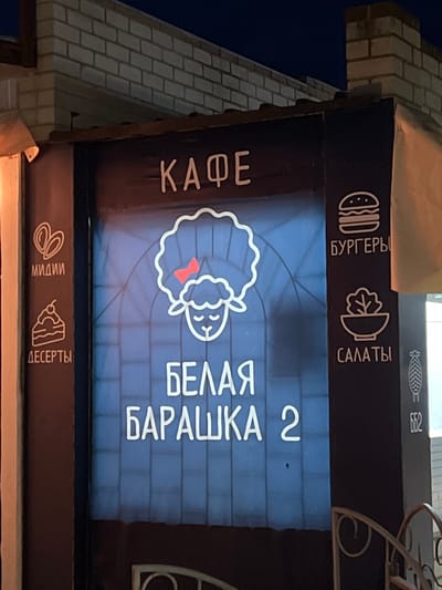 This media file captures a close-up, vertically oriented view of a sign for a cafe. The sign is illuminated and features various icons and text in Cyrillic.
At the top of the sign, there are three icons: a stylized burger, a salad bowl, and what appears to be a fishbone or intricate design. Accompanying these are the words "БУРГЕРЫ" (Burgers) and "САЛАТЫ" (Salads).
The main body of the sign displays a large, white outline of a sheep's head with a red bow tie. To the right of the sheep, large white Cyrillic lettering reads "БЕЛАЯ БАРАШКА 2" (White Lamb 2). To the left of the sheep, vertically oriented white Cyrillic text reads "КАФЕ" (Cafe). The background of this section is a blue brick pattern.
Below the main section, another dark brown horizontal sign is visible. It features icons of what could be dough twists or pretzels and a slice of cake, with the Cyrillic words "МИДИИ" (Mussels) and "ДЕСЕРТЫ" (Desserts) below them.
The lighting suggests it is nighttime or dusk, as the sign is brightly illuminated against a darker background. The sign is attached to a building with a light-colored brick facade on the left. The overall impression is that of a brightly lit advertisement for a food establishment.