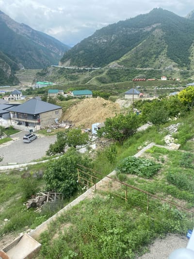 The image shows a residential area with mountains in the background. The structures visible include houses with gray and green roofs, and a few smaller buildings. A silver minivan is parked on a gravel driveway. The terrain is sloped with overgrown vegetation, including trees and garden plots. In the foreground, there is a low metal fence and some piled lumber. The sky is overcast. The scene depicts a rural or semi-rural environment.