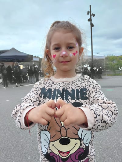 A young girl with face paint stands outside in what appears to be a parking lot or public area. She is wearing a patterned sweatshirt featuring a Minnie Mouse design. The face paint consists of hearts and other designs on her cheeks and nose. The background shows a scene with people gathered near a blue tent, along with streetlights and buildings in the distance. It is not possible to definitively determine the location as Taastrup, Denmark from this image alone. More information is required.