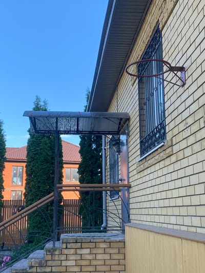 A photograph taken at a low angle shows the side of a light yellow brick house with a dark grey, ribbed roof overhang. A rusty basketball hoop is attached to the brick wall above a window with black wrought-iron bars. To the left, a covered porch with a patterned translucent roof is supported by dark posts. A metal railing with wooden accents leads up a brick staircase. Several tall, green cypress trees are planted in front of the house and a brown-tiled roof house is visible behind them. The sky is clear and blue. The location is Volzhsk, Russia.
