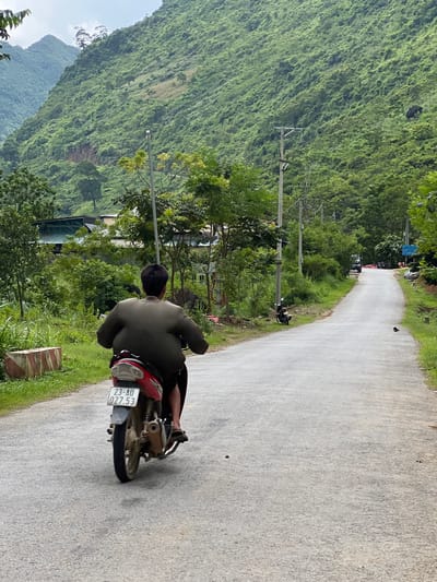 The image depicts a person riding a red motorcycle on a paved road in a rural area, likely Cán Tỷ Commune, Vietnam. The rider, seen from behind, is wearing a dark olive-green jacket and dark pants. They appear to have a large build, causing their jacket to bulge outwards significantly. The motorcycle has a license plate that reads "23-AU 027.53".
The road is asphalt and curves slightly to the right, leading into the distance. On either side of the road, lush green vegetation and trees are abundant. In the background, steep, heavily vegetated mountains dominate the landscape, with some structures, possibly houses or small buildings, visible amongst the trees on the lower slopes. Power lines and utility poles are also present along the side of the road. The sky is overcast. A small black bird is visible on the road in the middle ground, and other motorcycles are parked further down the road.