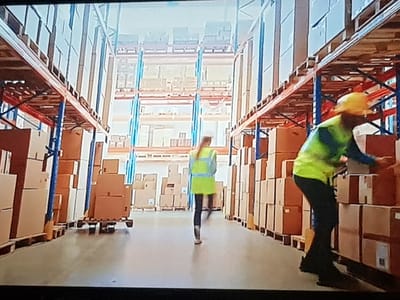 The image shows the interior of a large warehouse with tall shelves stacked with cardboard boxes. Two individuals are visible. One person, wearing a yellow hard hat and a bright green safety vest over a blue shirt, is in the foreground on the right, appearing to handle or examine some boxes. This person is wearing dark pants and shoes. The other person, wearing a similar bright green safety vest over a blue shirt and dark pants, is in the middle ground on the left, walking away from the camera. This individual also appears to be wearing a safety vest. The warehouse environment is characterized by rows of shelving units with metal supports and wooden planks holding the boxes. The floor is a smooth, light-colored concrete. The lighting suggests an indoor environment, likely with artificial lighting, creating a bright atmosphere. The specific location in Lomets, Bulgaria, cannot be determined from the visual information provided in the image.