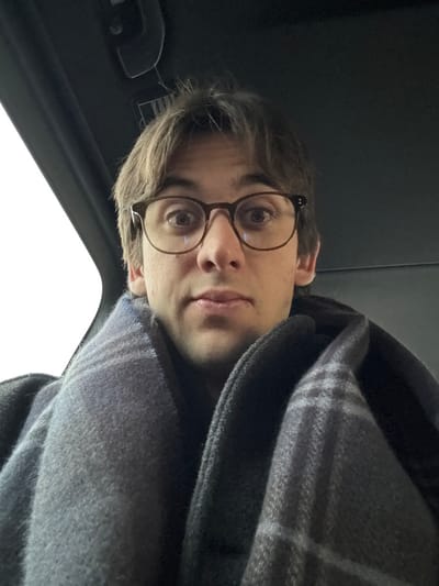 A male individual with brown hair and brown-framed eyeglasses is depicted in a close-up, upward-angled selfie taken inside a vehicle in Roissy-en-France, France. He has a light complexion, his eyes are wide, and his lips are slightly parted. He is wrapped in a grey and dark blue plaid-patterned blanket or scarf, visible around his neck and shoulders, over a dark garment. The vehicle's dark interior ceiling is visible in the upper portion of the image. A bright, featureless white light source, presumably an exterior sky, is visible through a window on the left side of the frame.