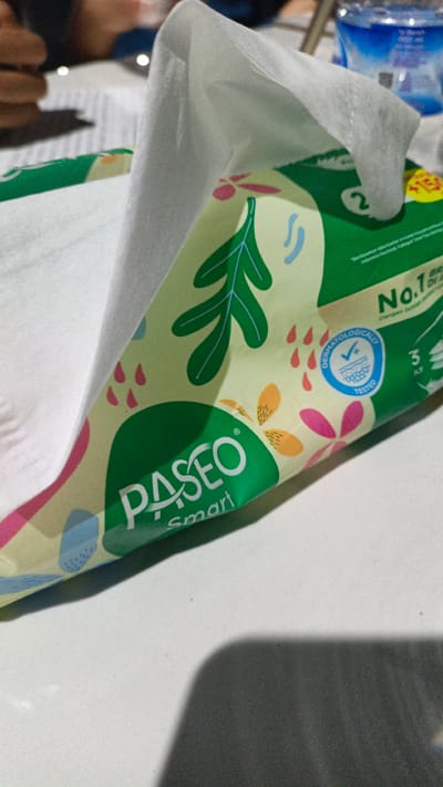 The image shows a close-up view of a package of Paseo smart facial tissues, likely in a home setting given the presence of what appears to be a table and hints of people in the background. The packaging has a light green and white design with colorful floral and leaf motifs. A white tissue is partially pulled out from the opening, suggesting the product is in use.

Prominently displayed on the packaging are the brand name "PASEO" in large green letters and the word "smart" in smaller, white text underneath. There are also various graphical elements and text, including a blue circular badge with "DERMATOLOGICALLY TESTED" and a checkmark inside. Other visible text includes "No.1" and "BR / DI" on the right side of the package, and "3 / LY". The overall impression is that of a personal care product being presented, with an emphasis on its dermatological testing and brand recognition. The lighting suggests it is daytime, possibly indoors. The scene is intimate and focused on the product itself, with the background softly blurred.