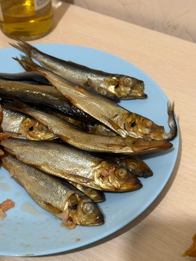 A light blue plate filled with smoked sprat fish is presented on a wooden table. The fish are arranged in layers, with their heads and tails clearly visible. The plate is slightly off-center to the right. In the upper left corner, a glass bottle containing a yellowish liquid, possibly beer or oil, is partially visible. The background consists of a plain wooden surface and a light-colored wall. The lighting is moderate, casting soft shadows. There are no people or discernible animals present in the scene. The image does not contain any visual elements that would indicate a specific location within the city of Ivano-Frankivsk, Ukraine.