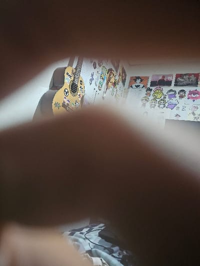 A sticker-covered guitar lies on a white surface, amid scattered stickers and papers. Anime-themed images adorn the wall behind, adding to the chaotic yet personalized atmosphere. The image is obscured by blurry foreground elements, possibly fingers, directing focus towards the central guitar and surrounding paraphernalia. A partial view of patterned fabric is visible on the right.