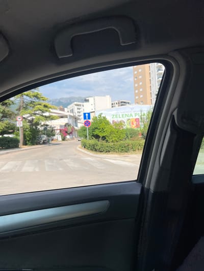 This image appears to be taken from inside a vehicle, looking out through a slightly open window or sunroof. The perspective is from a car, as evidenced by the interior of the car visible in the foreground: a dark grey interior panel with a grab handle.
The scene outside is an urban or semi-urban environment. A road with a pedestrian crossing is visible, along with buildings, trees, and foliage. One building is a tall, light-colored structure with multiple windows. There's a large billboard or advertisement visible, featuring green text that reads "DOO KOMUNALNE DJELATNOSTI" and "ZELENA PI". The colors on the billboard are vibrant, suggesting it depicts produce or a natural theme.
There are traffic signs visible: a blue sign with white arrows indicating direction, and a red circular sign with a diagonal line, likely a prohibition sign. Further down the road, another red circular sign with a number inside and a diagonal line is visible, potentially a speed limit sign. The presence of these signs and the road infrastructure suggest a developed area.
Lush green trees and bushes are abundant, with some pink flowers adding color to the scene. In the background, mountains are visible, partially obscured by the buildings and trees, indicating a mountainous or hilly terrain surrounding the location. The sky is a light blue with some scattered clouds, suggesting it is daytime and the weather is likely fair.
The image is captured at an angle, rotated roughly 90 degrees clockwise, making the landscape appear vertical. The overall impression is one of a bright, sunny day in a visually appealing area with a mix of urban development and natural greenery. The location context provided is Bjeliši, Montenegro, which aligns with the presence of mountains and potentially the architectural style of the buildings. No people are clearly visible in the scene.