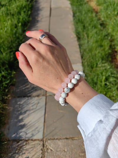 A close-up, medium shot captures a woman's hand and arm adorned with two beaded bracelets, a ring, and vibrant orange nail polish. She is wearing a white linen shirt, the collar neatly buttoned. The background consists of a blurred, sun-drenched outdoor setting with green grass and a paved walkway. The scene is illuminated by natural light, casting shadows. There are no discernible city landmarks or specific indicators to place the location within Fanipal, Belarus.