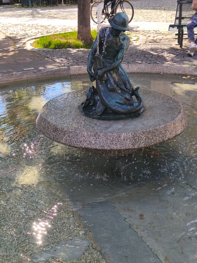 The image shows a bronze statue of a merman holding a fish, positioned in a fountain. Water cascades from the statue's head, creating ripples in the shallow pool. The fountain is surrounded by a cobblestone and paved area, with a grassy patch to the left and trees in the background. A bicycle and a park bench with a person sitting on it are visible in the distance, suggesting a public park or square setting in Erlangen, Germany. The sun is shining, casting reflections on the water.