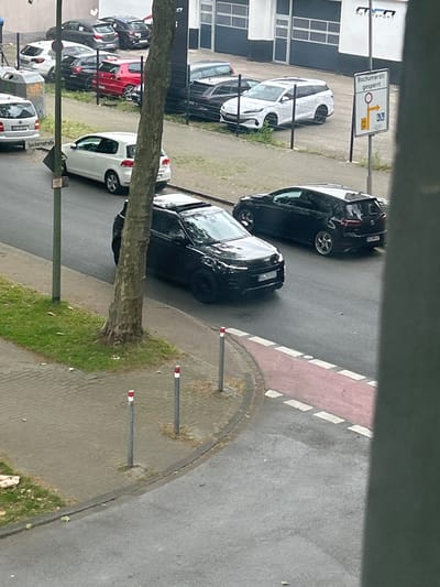 The image depicts a street scene in Gelsenkirchen, Germany. In the foreground, a dark-colored car is parked on the asphalt road, partially obscuring the view of a tree trunk. Behind the tree, another dark-colored car is driving on the road. Further down the street, a white station wagon is parked. To the right of the white station wagon, a white sign with text and symbols is mounted on a pole. This sign appears to be a traffic sign indicating a road closure and a detour. Several other cars are visible in the background, parked along the street and near a building that seems to be a commercial establishment, possibly a car dealership or repair shop, given the "MOTORSPORT" signage. The area includes a sidewalk, grass, and bollards along the edge of the sidewalk. The overall impression is a typical urban street environment.