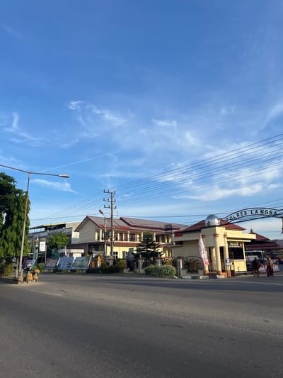 The image depicts the entrance to POLRES LANGSA in the city of Langsa, Indonesia, on a clear day with a blue sky and scattered white clouds. A bright sun casts shadows, indicating either morning or late afternoon.

The POLRES LANGSA building is a prominent yellow structure with a red-tiled roof, featuring a small domed section above the main entrance. A sign above the entrance arch reads "POLRES LANGSA". To the left of the entrance is a security post labeled "PENJAGAAN".

In front of the building, there are several banners displayed, some featuring graphics and text. A large green tree stands to the left of the road, partially obscuring a streetlight. Power lines and utility poles are visible running across the top of the frame.

On the road in front of the POLRES building, two individuals are walking away from the camera towards the right. They are dressed in traditional clothing, with one person wearing a brown garment and the other in a beige headscarf and dress. A white bus with "POLISI" written on its side is parked in the background.

The foreground shows a paved road, with the right lane appearing slightly darker than the left. The overall scene suggests a calm, everyday setting in front of a government facility in Langsa.